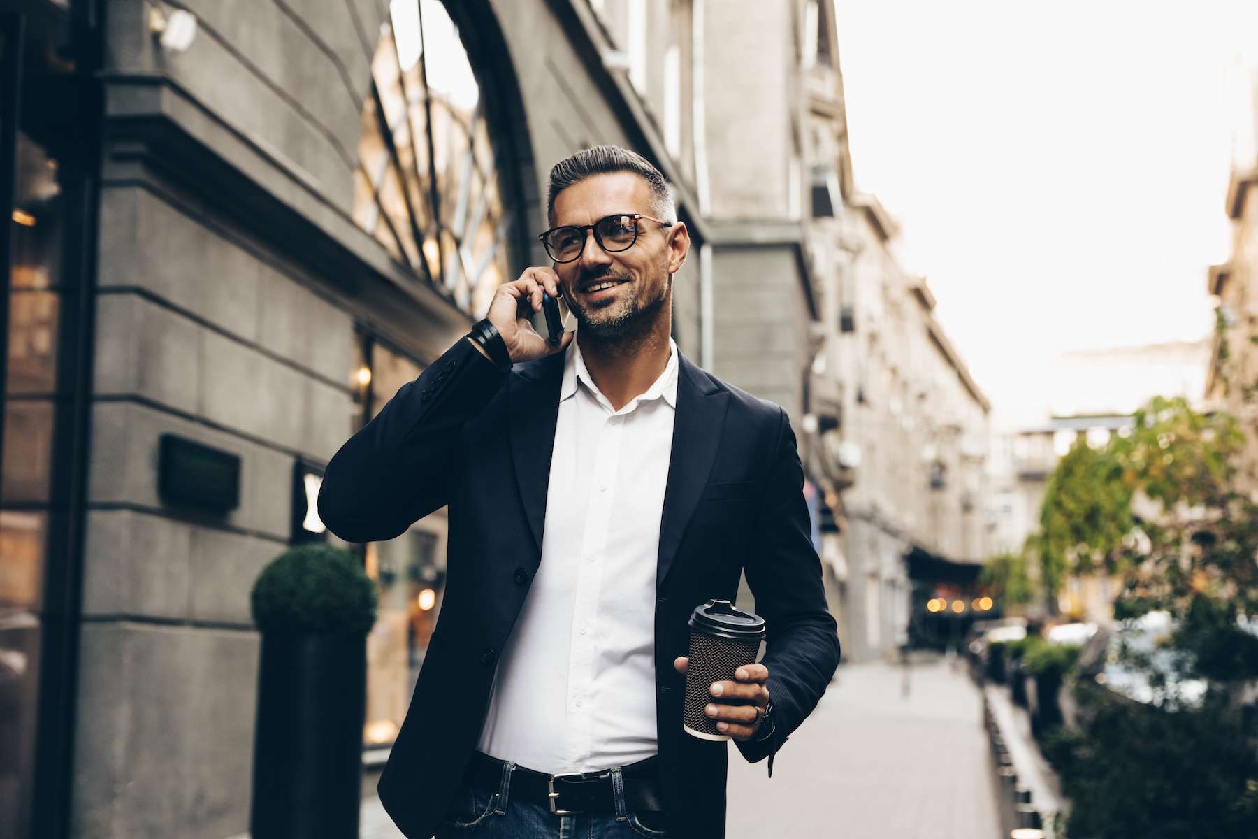 mature male walking through city sidewalk, talking on the phone, holding a cup of coffee